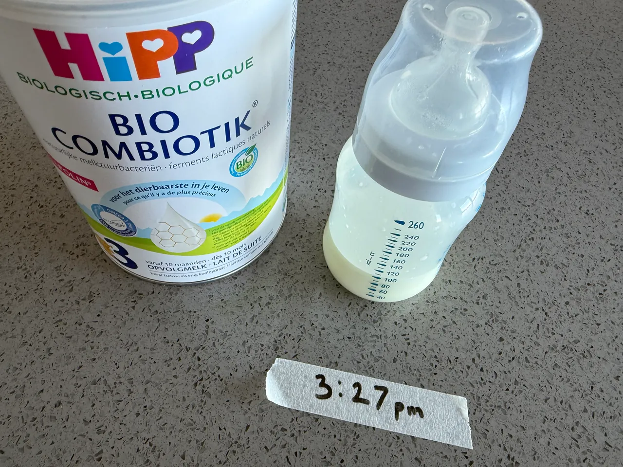 A made-up bottle of formula beside a tin of HiPP Bio Combiotik Stage 3, and in front of them a strip of masking tape with the time the bottle was made written on it.
