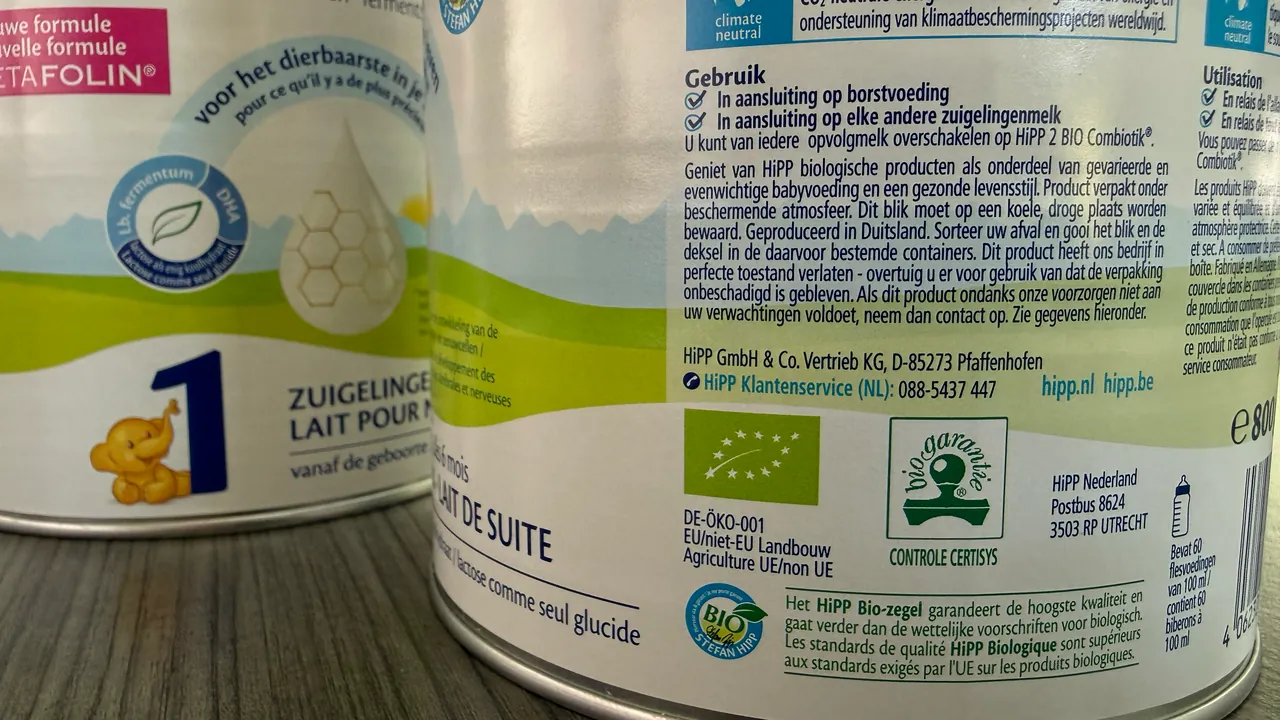 Two HiPP Dutch formula tins on a wooden table: the front of the Stage 1 tin on the left, and on the right the back of the Stage 2 tin, which names HiPP GmbH & Co. Vertrieb KG in Pfaffenhofen, Germany, and HiPP Nederland in Utrecht, beside the EU organic leaf.