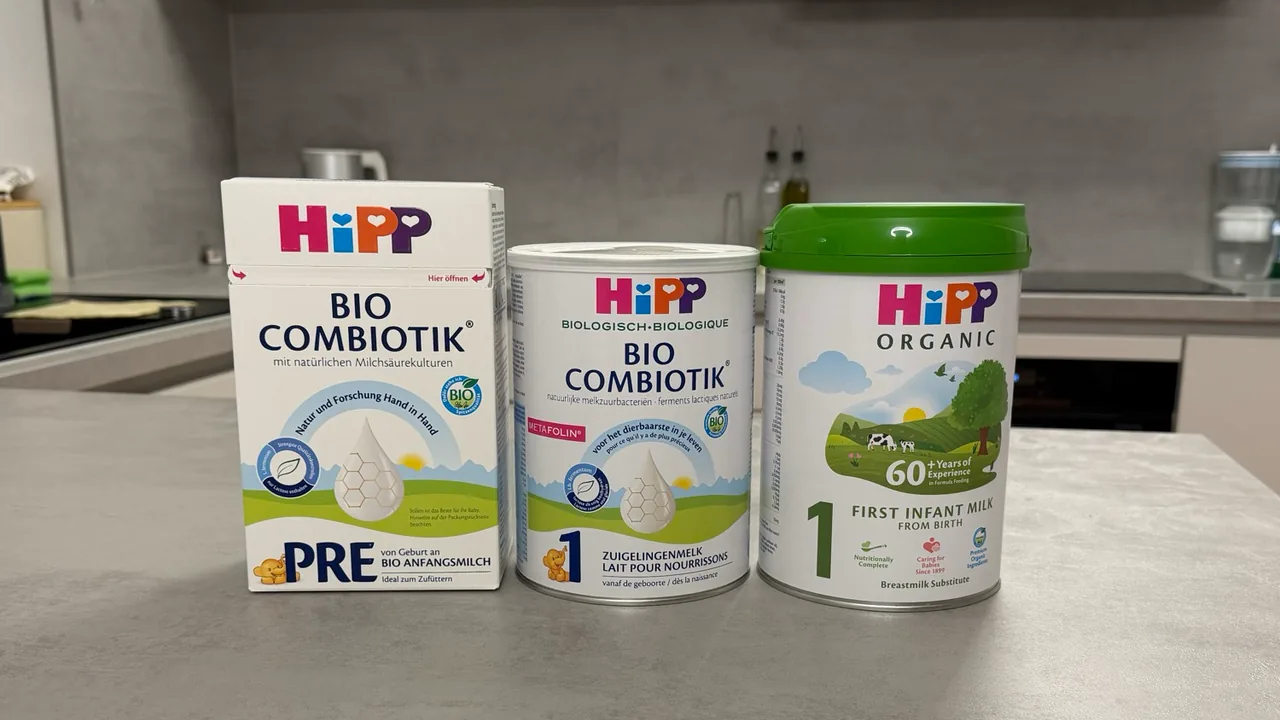 Three HiPP formulas side by side on a kitchen counter, fronts to the camera: the German PRE box, the Dutch Stage 1 tin and the UK First Infant Milk tin.