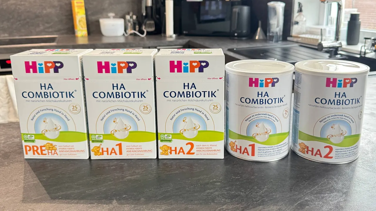 Five HiPP HA Combiotik packs in a row on a kitchen counter, fronts to the camera: the German HA PRE, HA 1 and HA 2 boxes, then the Dutch HA 1 and HA 2 tins.