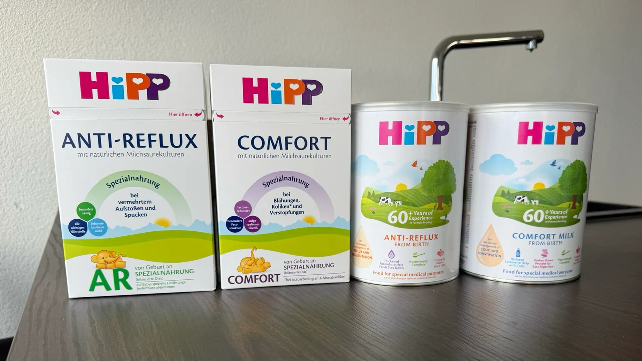 Four HiPP packs on a kitchen counter, fronts to the camera: the German Anti-Reflux and Comfort boxes, and the British Anti-Reflux and Comfort Milk tins.
