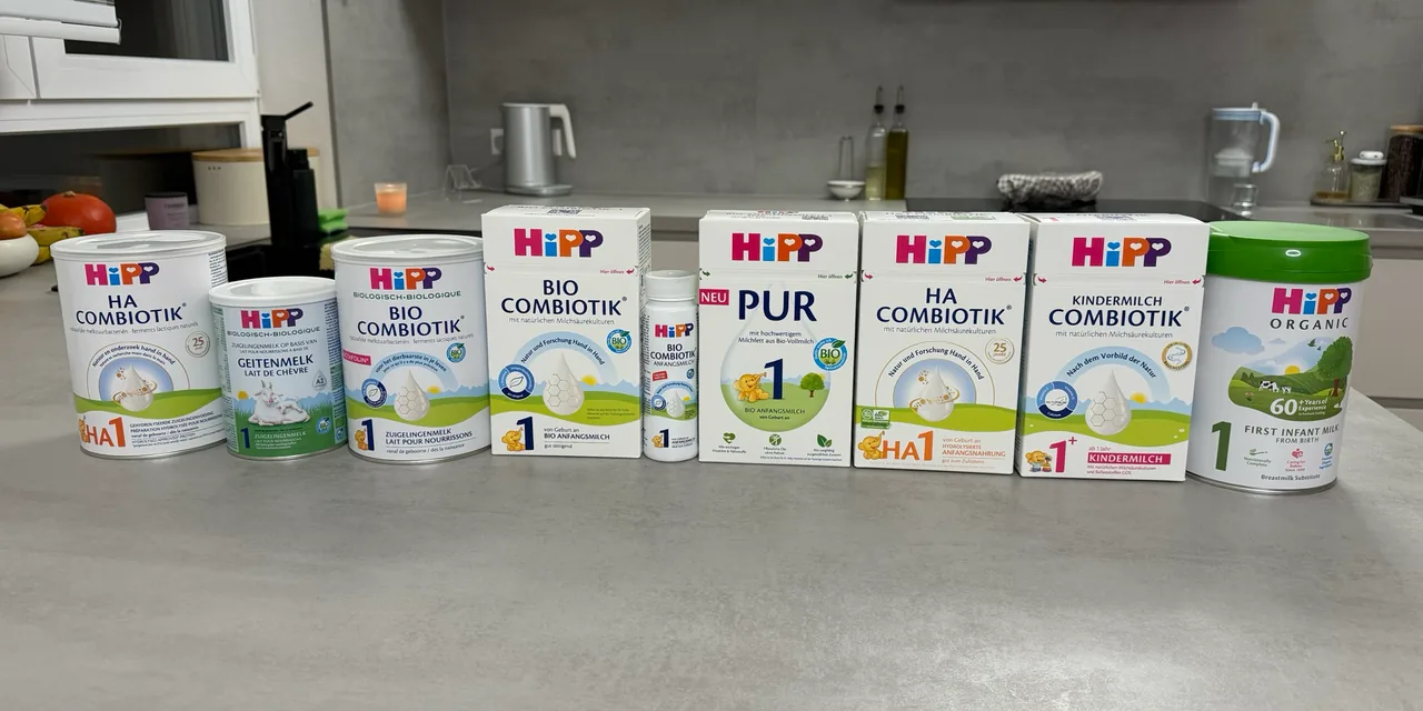 Nine HiPP packs in one row on a kitchen counter, fronts to the camera: the Dutch HA 1, goat's milk 1 and Stage 1 tins, the German Stage 1 box and its ready-to-feed bottle, the PUR 1 and German HA 1 boxes, a Kindermilch 1+ box and the UK First Infant Milk tin.