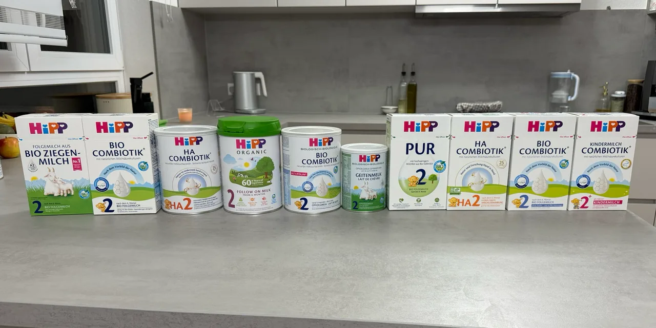 Ten HiPP packs in one row on a kitchen counter, fronts to the camera: the German goat's milk 2 and Stage 2 boxes, the Dutch HA 2 tin, the UK Follow On Milk tin, the Dutch Stage 2 and goat's milk 2 tins, the PUR 2, German HA 2 and German Stage 2 No Starch boxes, and a Kindermilch 2+ box.