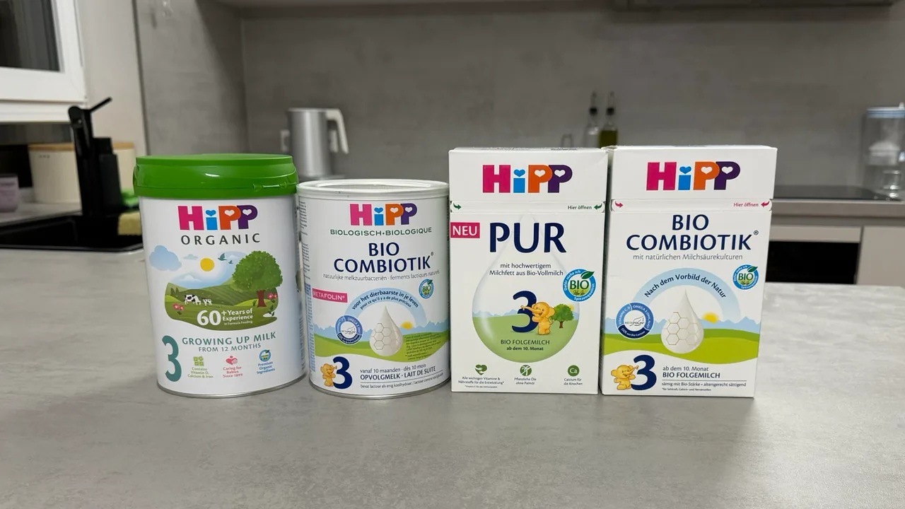 Four HiPP Stage 3 packs in a row on a kitchen counter, fronts to the camera: the UK Growing Up Milk tin, the Dutch Stage 3 tin, and the PUR 3 and German Stage 3 boxes.