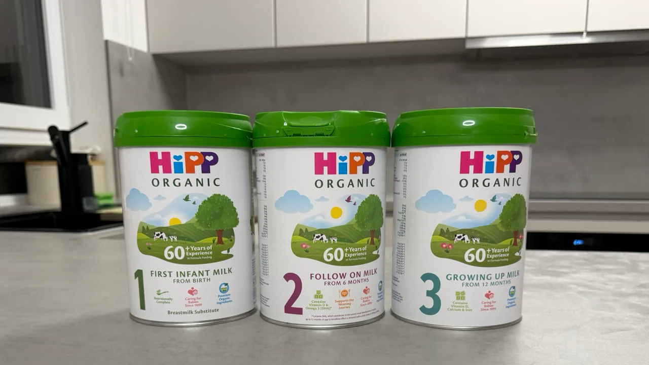 Three HiPP Organic tins from the UK in a row on a kitchen counter, fronts to the camera: First Infant Milk, Follow On Milk and Growing Up Milk.