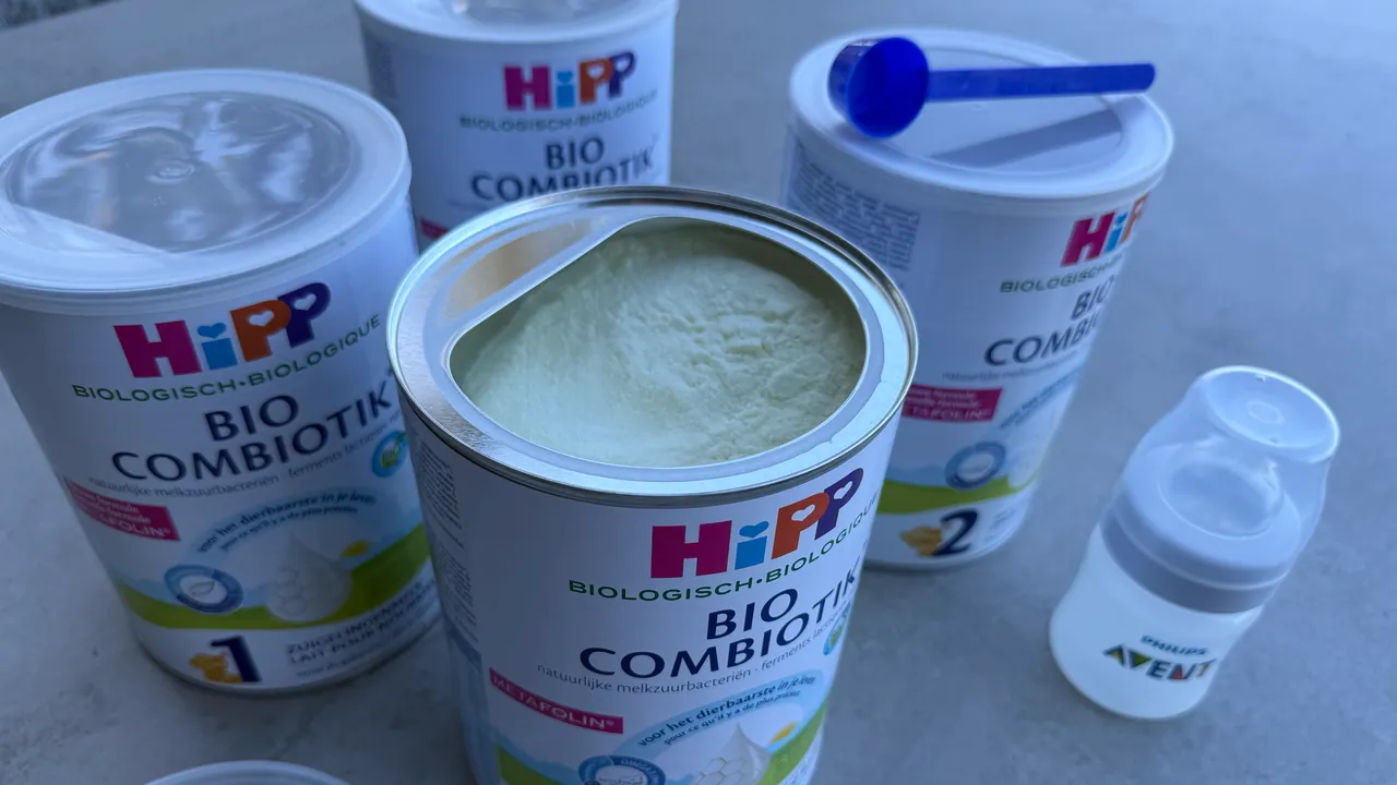 Cuatro latas de HiPP Bio Combiotik, la fórmula holandesa, sobre una encimera gris: una abierta para mostrar el polvo, una cuchara medidora azul sobre la tapa de otra, y un pequeño biberón junto a ellas.