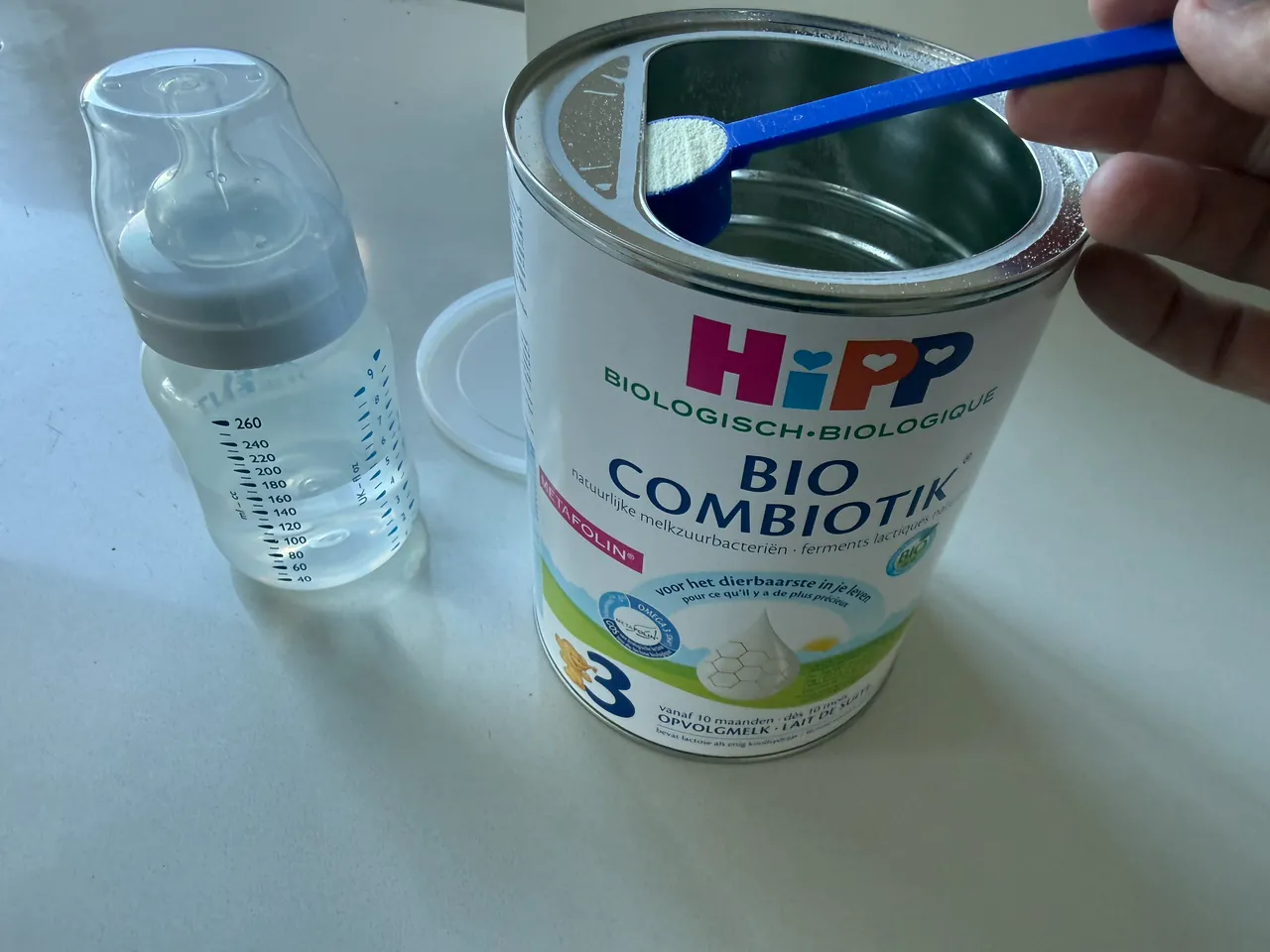 A hand holding a level scoop of powder against the flat edge inside an open tin of HiPP Bio Combiotik Stage 3, with a feeding bottle of water beside it.