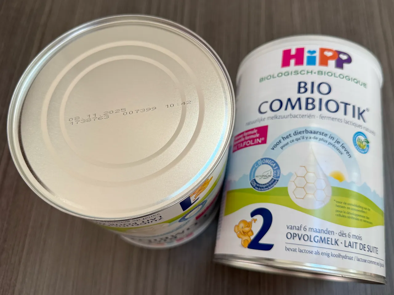 Two HiPP Bio Combiotik tins on a wooden table: one upside down, its base printed in small dots with a date and the batch details, and beside it the front of a Stage 2 tin.