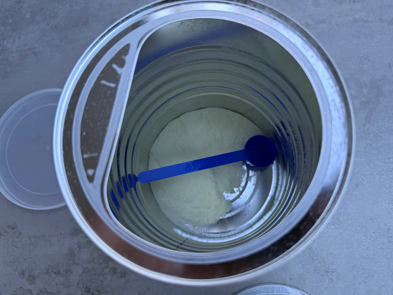 Looking down into an opened HiPP tin with little powder left, its blue scoop lying inside and the clear plastic lid set to one side.