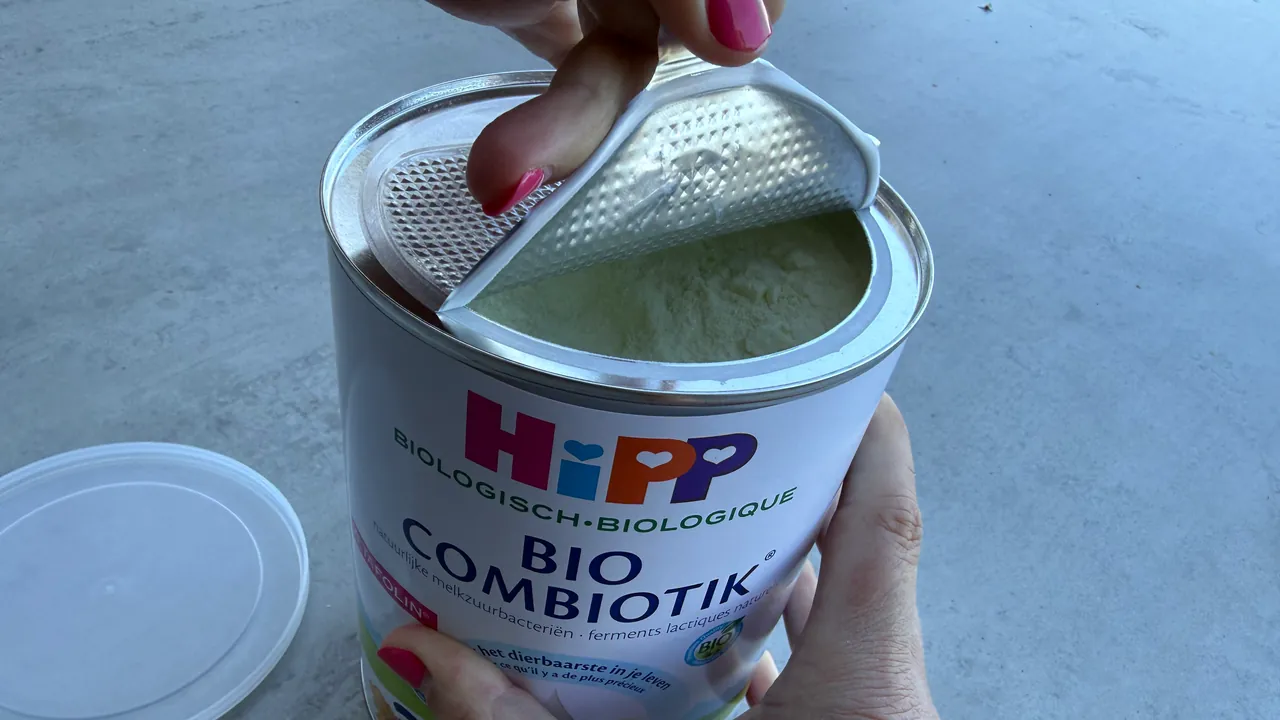 Hands peeling the foil seal back from a new tin of HiPP Bio Combiotik, the powder showing underneath, with the clear plastic lid set aside on the counter.